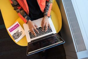 _Overhead view of woman’s hands typing at laptop, seated on yellow chair, working on coding file with a textbook beside her