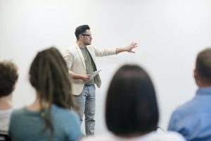 Clear view of professor teaching in the background with a row of blurry backs of heads of students in foreground learning