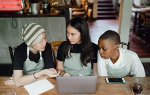 Face on view of three university students sitting in front of laptop in mid-discussion working on a project online