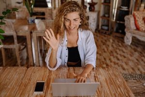 Happy woman sitting at table smiling, and waving at laptop while engaged in a video conference