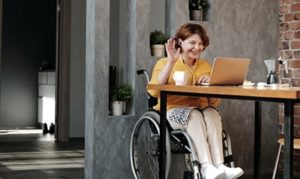 Woman in wheelchair sitting at table at home in front of open laptop, smiling and waving and chatting online