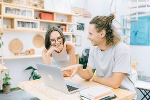 Young man and woman pointing and laughing sitting at table in at-home studio with books, pointing at open laptop