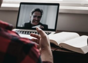 Over the shoulder view of a man in front of laptop with an open textbook, video conferencing with professor in the middle of an enthusiastic conversation
