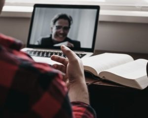 Over the shoulder view of man at desk with open textbook, motioning with his hand in a video conference on laptop with professor