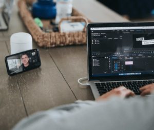 Widok przez ramię osoby pracującej na laptopie na biurku podczas wideoczatu na telefonie komórkowym na stojaku po lewej stronie laptopa