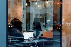 Through the window view of pensive woman in a communal workspace engaged in a video chat on smartphone leaning against open laptop