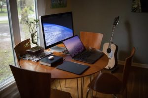 View of computer set up for at home learning including a desktop, laptop, and closed tablet on table with a ficus and guitar in background