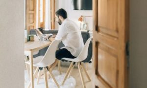 View of man through an open door in a modern living room working from home seated at desk on laptop