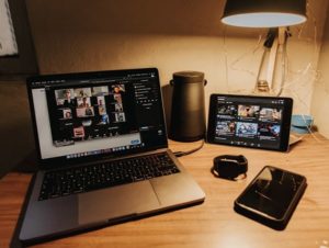 View of video conferencing gallery tiles on laptop beside speaker, tablet with music, watch and smartphone spread out on desk