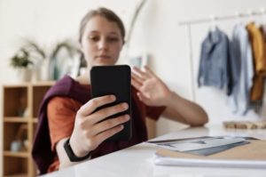 View of woman in midground using phone in foreground to join an online meeting. She is waving at her phone