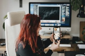 Woman sitting at desk in front of desktop working while holding up device and engaged with 4 speakers in a video conference