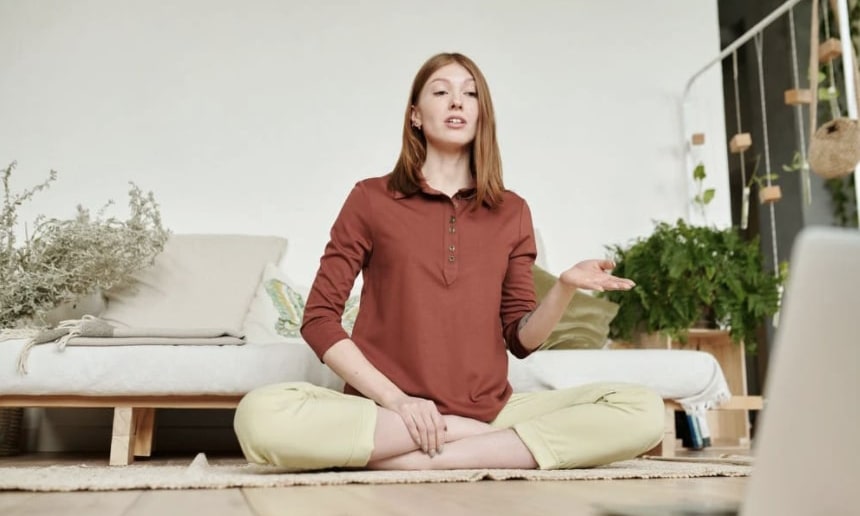 Yoga instructor in lotus position on ground interacting with laptop in front of her and leading a class