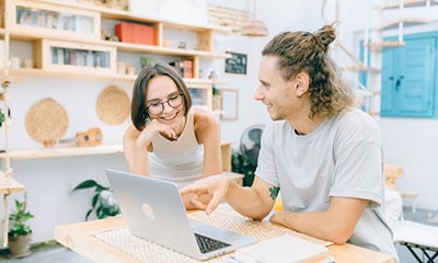 Jonge man en frou dy't wiisde en laitsje sitten oan tafel yn atelier thús mei boeken, wiisde nei iepen laptop-min