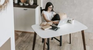 Midground view of young woman on laptop with textbooks studying at desk from home in bright and open loft space