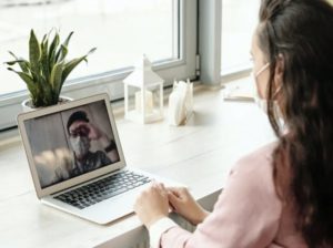 Por cima do ombro, uma mulher mascarada sentada em uma mesa de videoconferência com um homem mascarado em um laptop