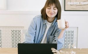 Straightforward view of smiling young woman drinking coffee while working on laptop at desk in communal working space