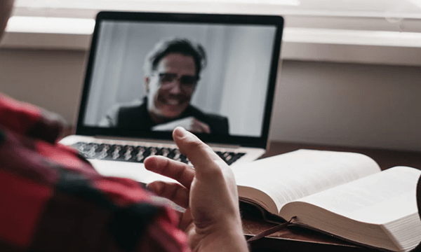 Over the shoulder view of man at desk with open textbook, motioning with his hand in a video conference on laptop with professor