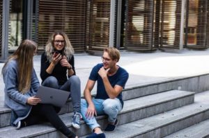 Vista de tres estudantes sorrintes comprometidos e charlando sentados nas escaleiras exteriores, interactuando cun portátil e un teléfono intelixentes abertos