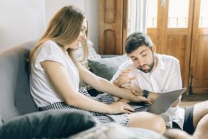 View of young woman sitting on couch and young man on floor, using his hand to gesticulate while interacting with open laptop at home