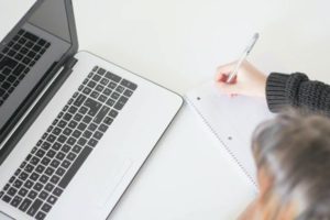 Angled view of woman writing in notebook while seated in front of open laptop on white surface