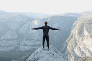 Backside view of inspired man standing on edge of a rock, arms uplifted facing a big and beautiful sunlit gorge