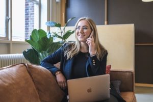 Happy, smiling woman seated on couch in sunny office while chatting on the phone with laptop open on her lap