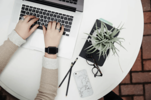 Overhead view of hands tapping on laptop beside smartphone, glasses, pencil and plant laid out on white round table