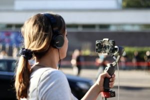 Side view of woman wearing headphones recording outdoors, holding mobile phone mounted to stabilizer