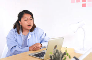 Young woman seated at desk facing laptop chatting to the open screen, at home in bright and white room