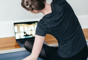 Young woman seated on yoga mat, midpose facing laptop on floor while engaged in a video conferencing yoga class