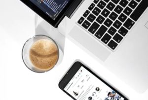 Bird’s eye view of corner of laptop, beside cappuccino and smartphone on white background