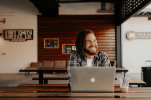 Casual-looking man with laptop, smiling and looking into the distance to the right, seated at a picnic bench in a coffee shop-min