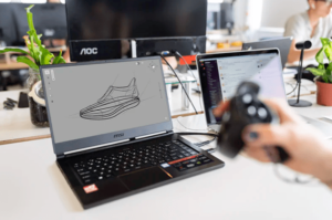 Focused view of open laptop in an office studio with digital sketch of shoe onscreen and blurred out hand holding a controller in foreground-min