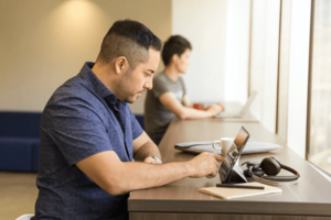 Man sitting at communal desk near window, engaged on laptop, using finger to navigate screen, with another person on laptop in background-min