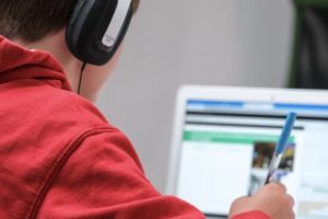 Over the shoulder view of a teen boy with headphones looking at laptop, pen in hand and writing in notepad