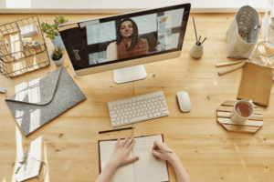 Pemandangan cerah dari meja kayu dengan cangkir kopi, tanaman, dan perlengkapan kantor; dua tangan menulis di notebook dan obrolan video di komputer desktop-min