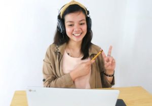 Young smiling woman seated at desk in front of laptop wearing headphones, teaching and communicating with hands against a white wall