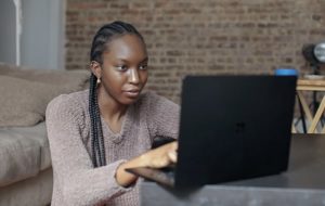 Angled view of a young woman using laptop on table, seated on ground at home beside couch with exposed brick in background