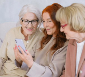 Direct view of three senior women seated together with middle woman holding a device and all three engaged, smiling and interacting with it-min