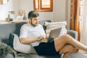 View of a casually dressed man at home working on laptop in reclined position on couch in brightly lit living room-min