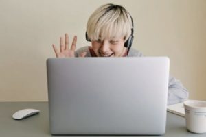 View of opened laptop partially covering body of young woman smiling and waving, seated at desk interacting with screen wearing headphones