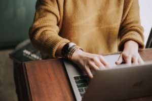 View of person from the neck down, wearing a mustard-colored sweater, sitting at a desk typing and interacting with a laptop