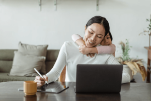 Sicht op frou mei in jong bern dy't har fan efter knuffelt, sit by buro thús yn wenkeamer op laptop mei in tablet-min