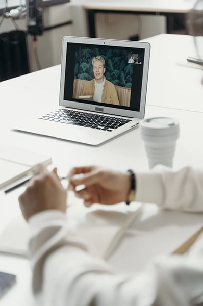 Kaffekop i forgrunden med åben bærbar computer på bordet i mødelokalet, der viser en ung mand videokonferencer set i billede-i-billede