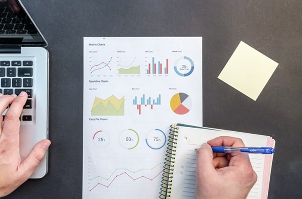 Overhead -skrivebord med en side med diagrammer og metrics, en klistermærke, den ene hånd skriver i en notesbog og den anden hånd ved hjælp af en bærbar computer