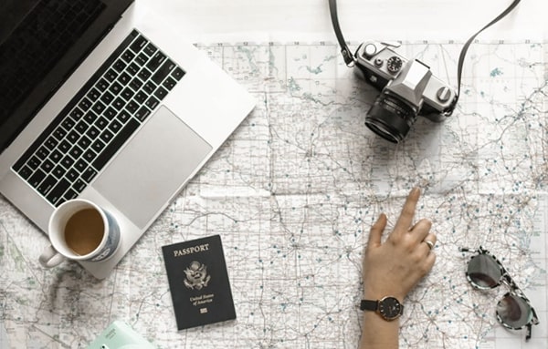 Overhead view of an opened laptop with passport, camera, and sunglasses laying on map with a finger pointing to specific location