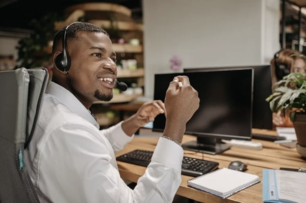 Side view of employee smiling and in mid-conversation, seated at desktop computer in office space with headphones, looking to the right