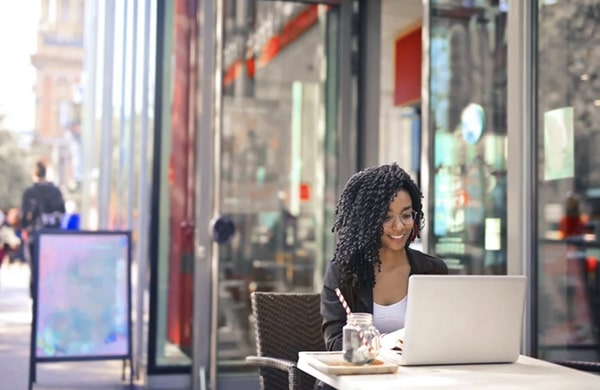 Smiling woman sitting at an outdoor table by the shop window, working on a laptop with a drink by her side