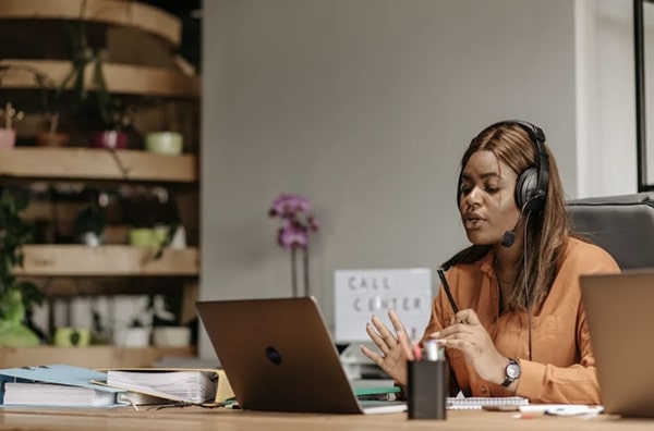 View of employee seated at desk in front of laptop wearing headphones in the middle of a call, gesticulating and speaking