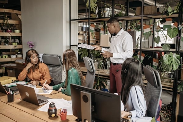 View of manager standing and meeting with three employees turned around listening to him delegate, while seated at desktop computers in office space